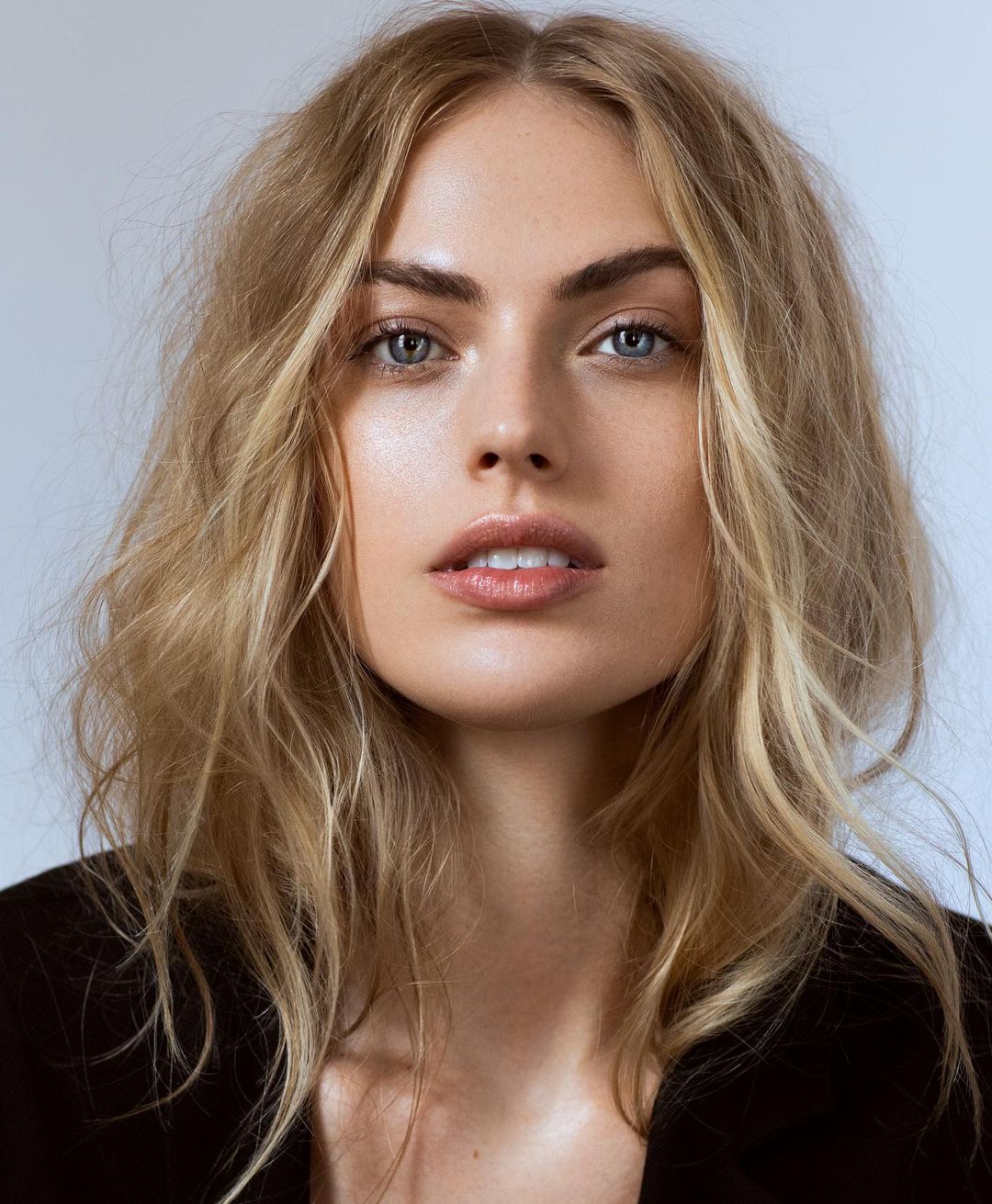 Close-up portrait of a woman with blonde hair, blue eyes, and natural makeup against a plain background
