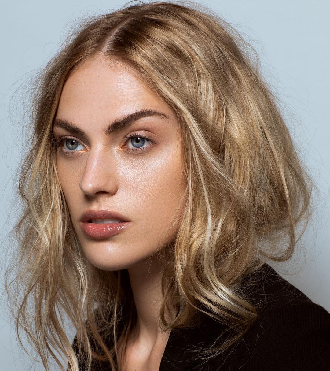 Close-up studio portrait of an adult female model with blonde wavy hair wearing a black blazer against a light blue background.