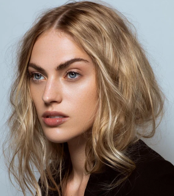 Close-up studio portrait of an adult female model with blonde wavy hair wearing a black blazer against a light blue background.
