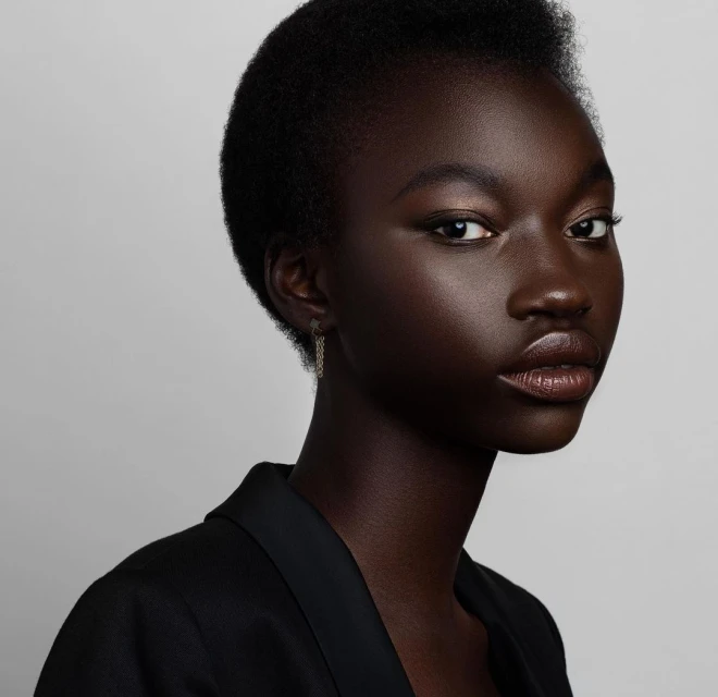 Close-up studio shot of a black adult female model with short hair wearing a black jacket against a light grey background, looking at the camera.