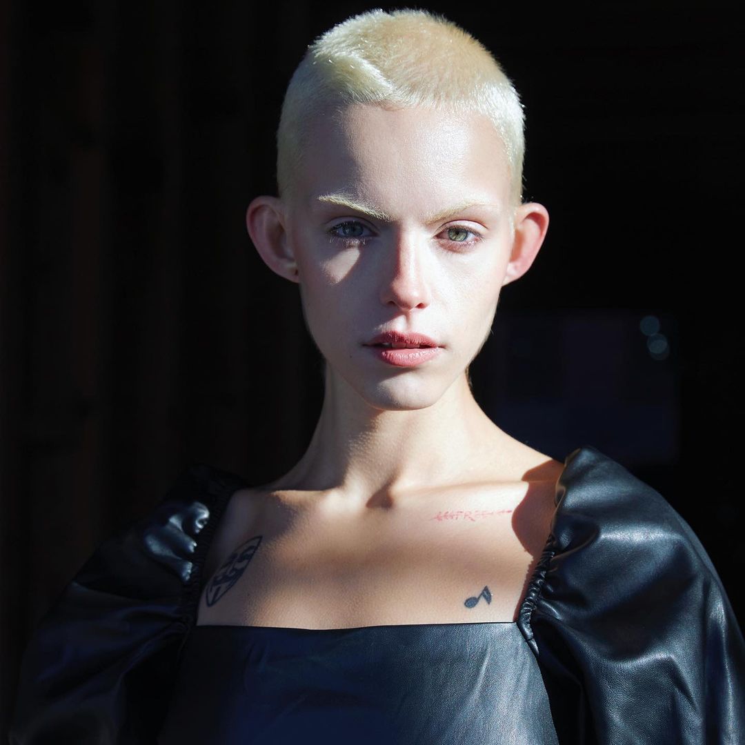 Close-up portrait of a fair-skinned, androgynous adult with a shaved head, wearing a black top, against a dark background.