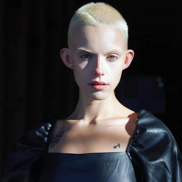 Close-up portrait of a fair-skinned, androgynous adult with a shaved head, wearing a black top, against a dark background.