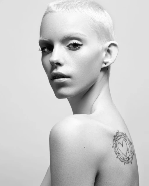 Portrait of a fair-skinned adult female with short blonde hair and a tattoo on her shoulder, set against a plain light background.