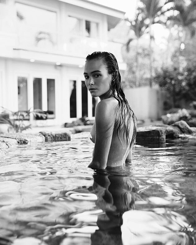 Monochrome portrait of a woman standing in water in front of a building surrounded by trees, creating a serene and natural atmosphere.