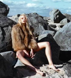 A blonde woman in a fluffy brown faux fur jacket seated among large coastal rocks on a sandy beach under a partly cloudy sky.