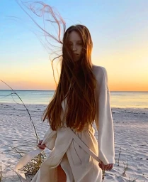 Woman with long brown hair blowing in the wind wearing a cream robe on a sandy beach during sunset.