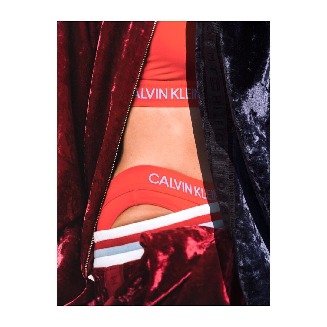 Close-up of a woman's torso wearing a red Calvin Klein sports bra and underwear set, partially covered by dark velvet jackets.