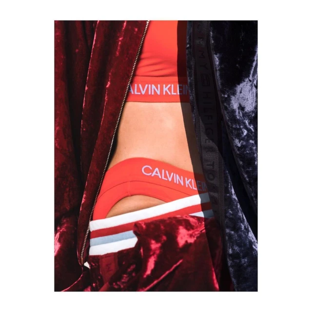 Close-up of a woman's torso wearing a red Calvin Klein sports bra and underwear set, partially covered by dark velvet jackets.