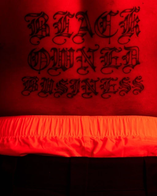 Close-up of a torso featuring a 'Black Owned Business' tattoo, with the lower part of the body partially visible in a vivid orange color.