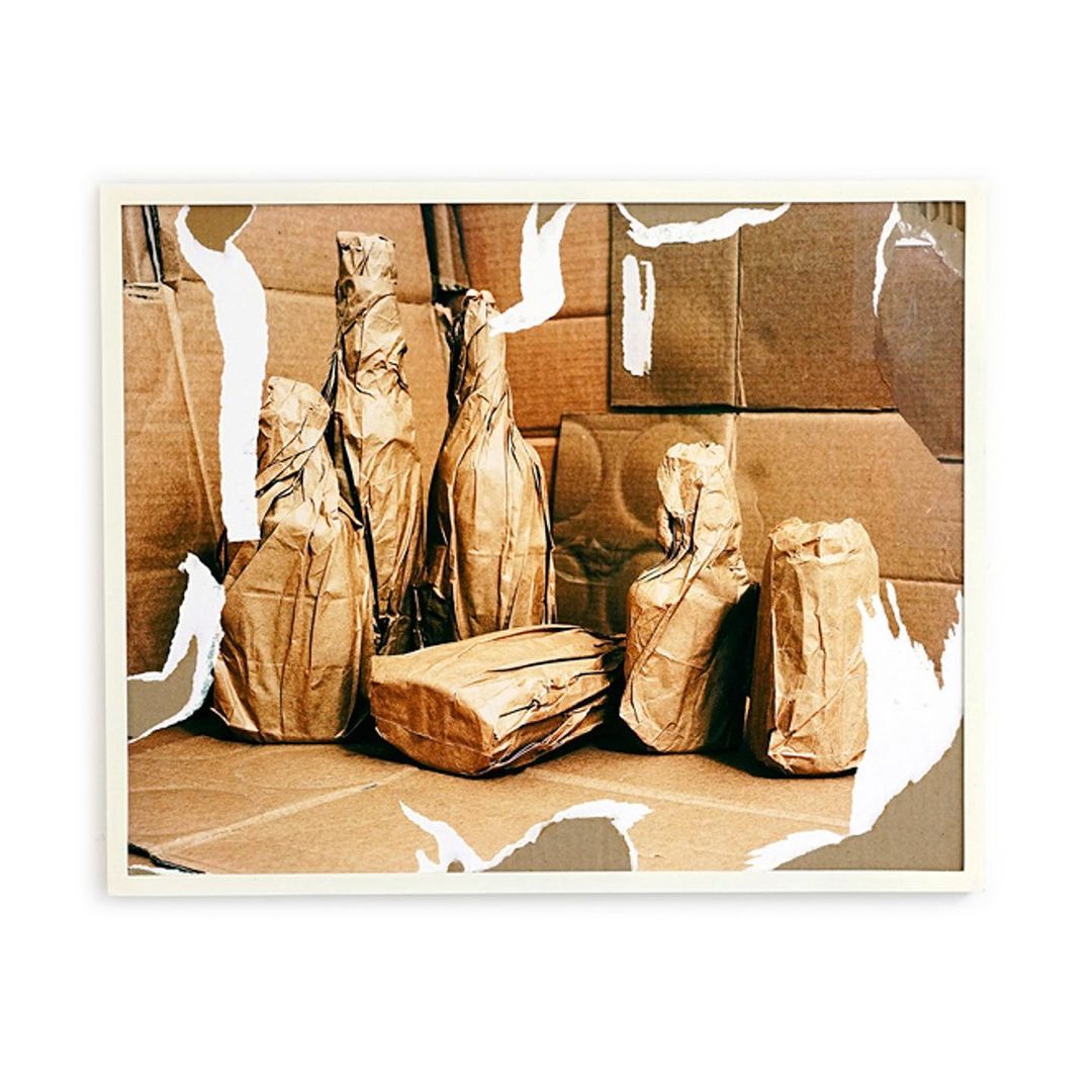 Still life photograph of brown paper-wrapped bottles and packages arranged on a cardboard surface, presented within a white frame.