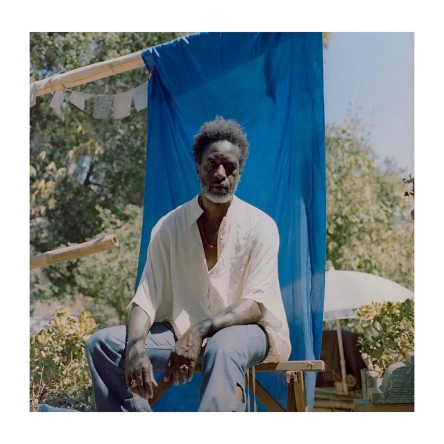 Portrait of a mature African-American man with a gray beard, seated outdoors with a blue fabric backdrop and trees in the background.