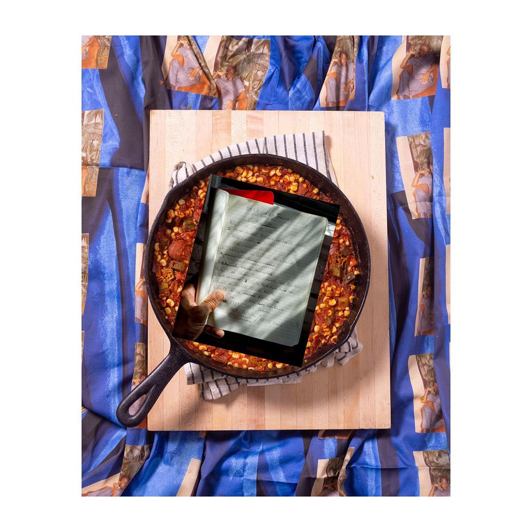 Overhead view of a cast-iron skillet with food on a wooden board, a hand holding a document, and a patterned blue tablecloth.