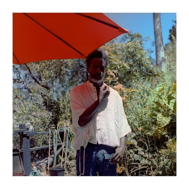 Outdoor portrait of an adult Black man with a beard under an orange umbrella, wearing a white button-down shirt in a garden setting.