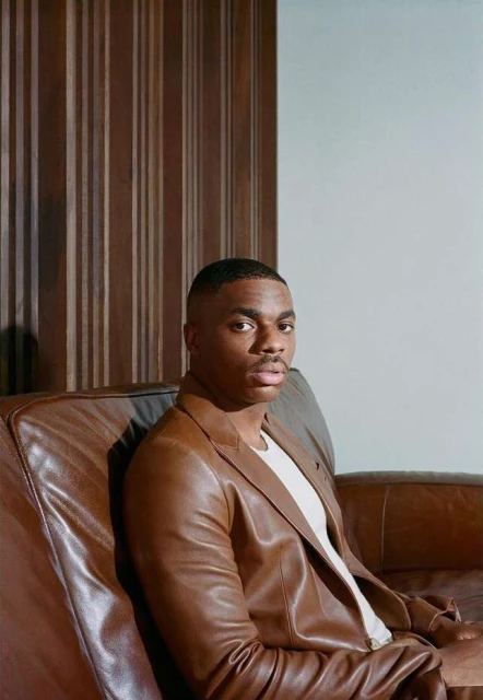 Stylish adult male portrait: Man with dark hair and brown jacket poses on leather couch in an indoor setting with neutral background.