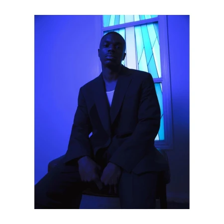 Portrait of a black man wearing a dark blazer sitting in front of a blue stained glass window