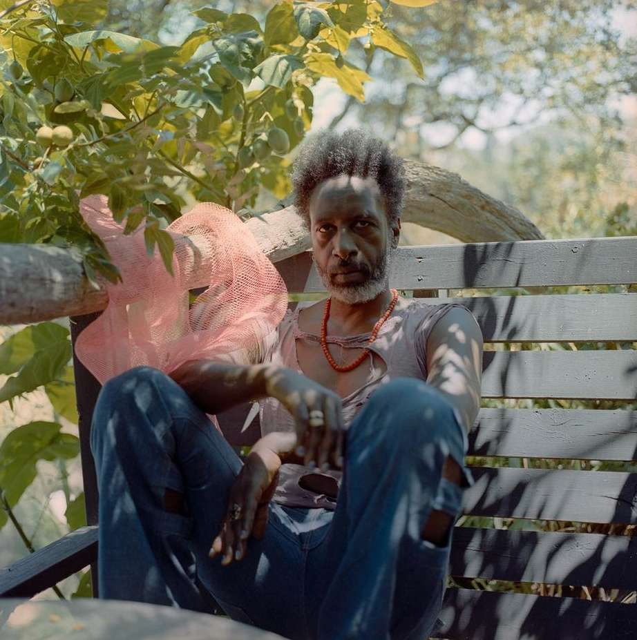 Adult Black man sits on a wooden bench outdoors, wearing jeans, a ripped t-shirt, and red beaded necklace, with foliage background.