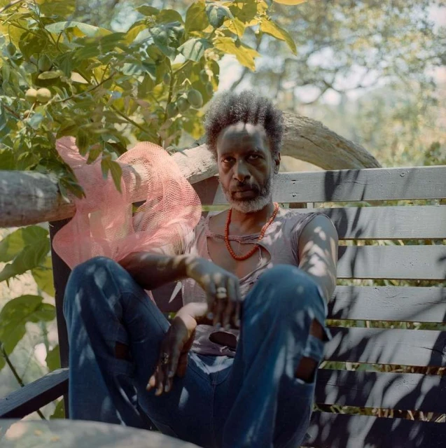 Adult Black man sits on a wooden bench outdoors, wearing jeans, a ripped t-shirt, and red beaded necklace, with foliage background.