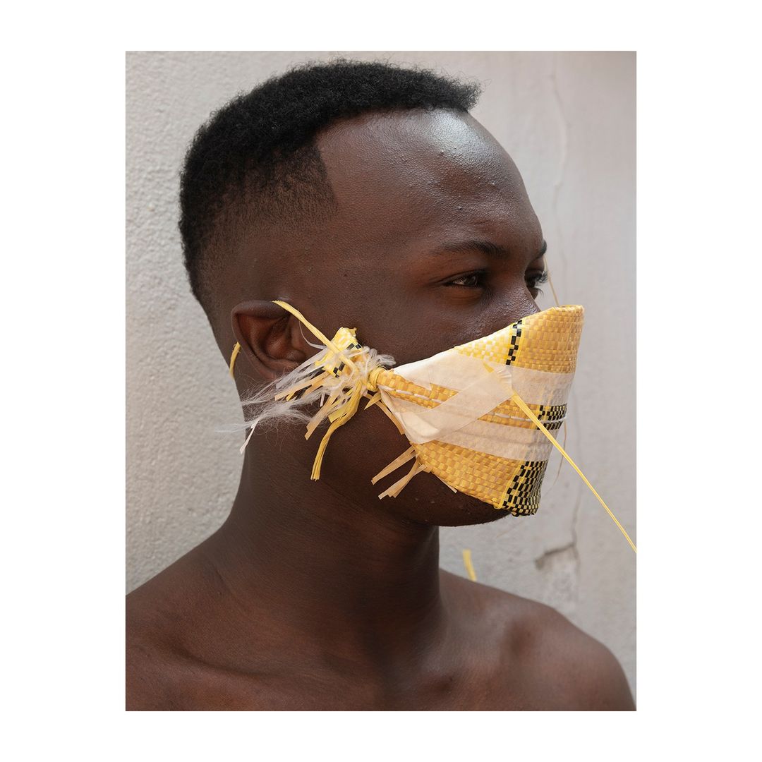 Close-up profile of an African man wearing a makeshift yellow and white mask with string ties, set against a plain light-colored wall.