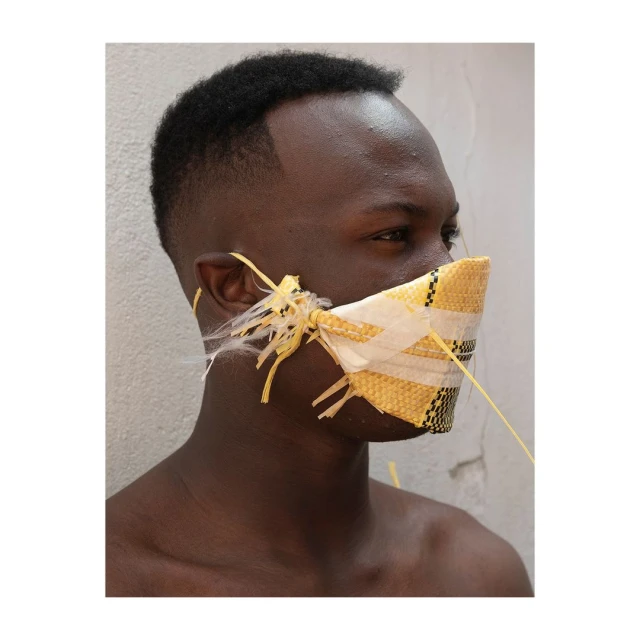 Close-up profile of an African man wearing a makeshift yellow and white mask with string ties, set against a plain light-colored wall.
