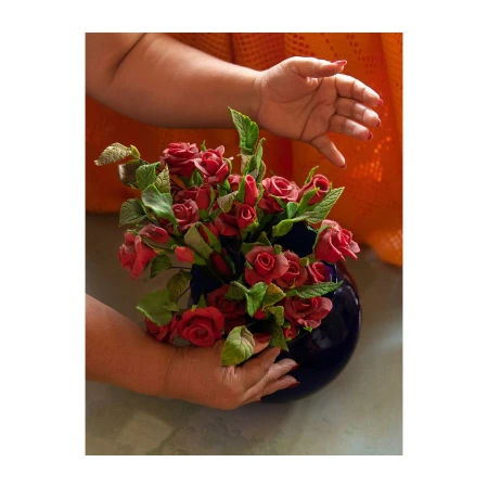 Close-up of hands arranging red clay flowers in a dark blue vase against a textured orange backdrop