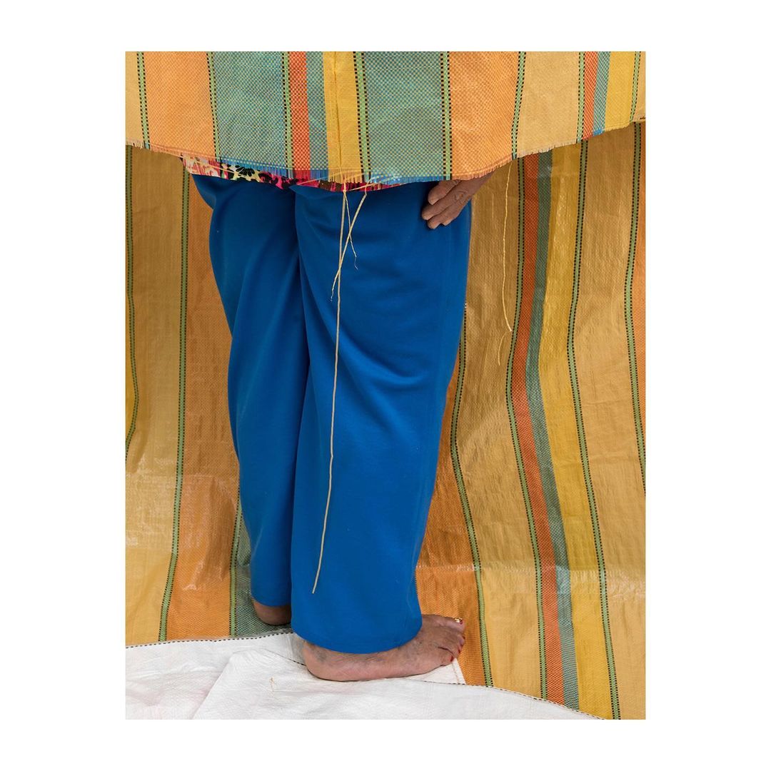 A person stands with bare feet in blue pants against a tan, striped backdrop with a colorful striped fabric above.