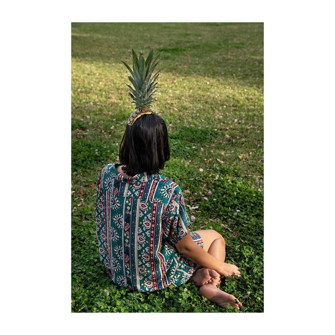 Young woman with patterned shirt sitting on a lawn with a pineapple on her head, back view, sunny outdoor setting.