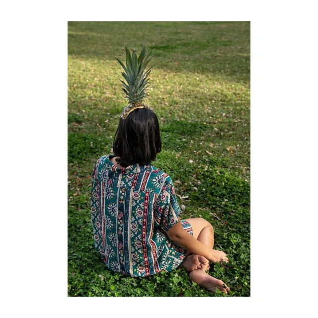 Young woman with patterned shirt sitting on a lawn with a pineapple on her head, back view, sunny outdoor setting.