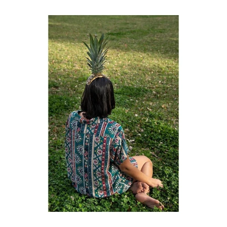 Young woman with patterned shirt sitting on a lawn with a pineapple on her head, back view, sunny outdoor setting.