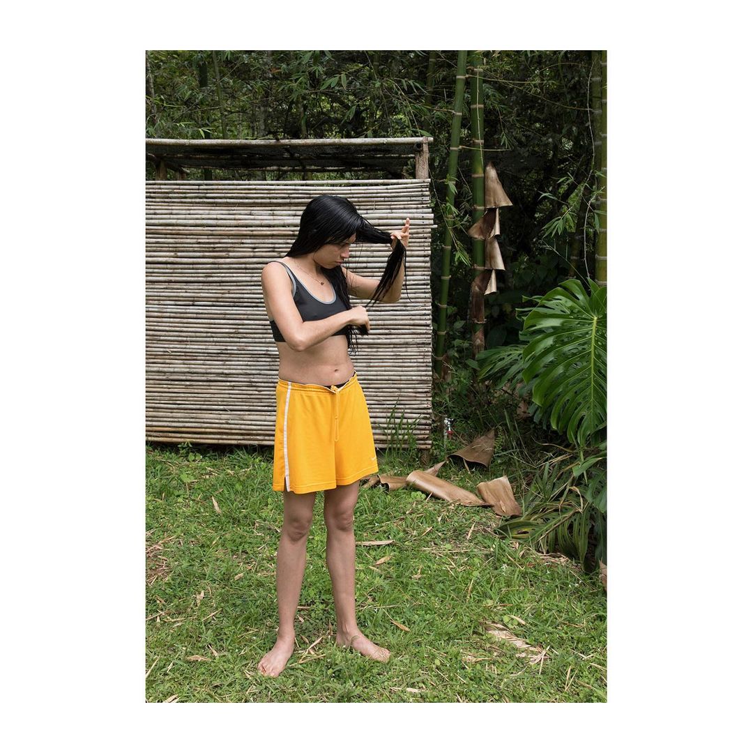 Young woman with long dark hair in swimwear and shorts stands in a grassy outdoor setting, grooming her hair near a bamboo structure.