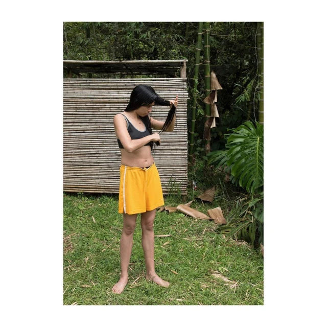 Young woman with long dark hair in swimwear and shorts stands in a grassy outdoor setting, grooming her hair near a bamboo structure.