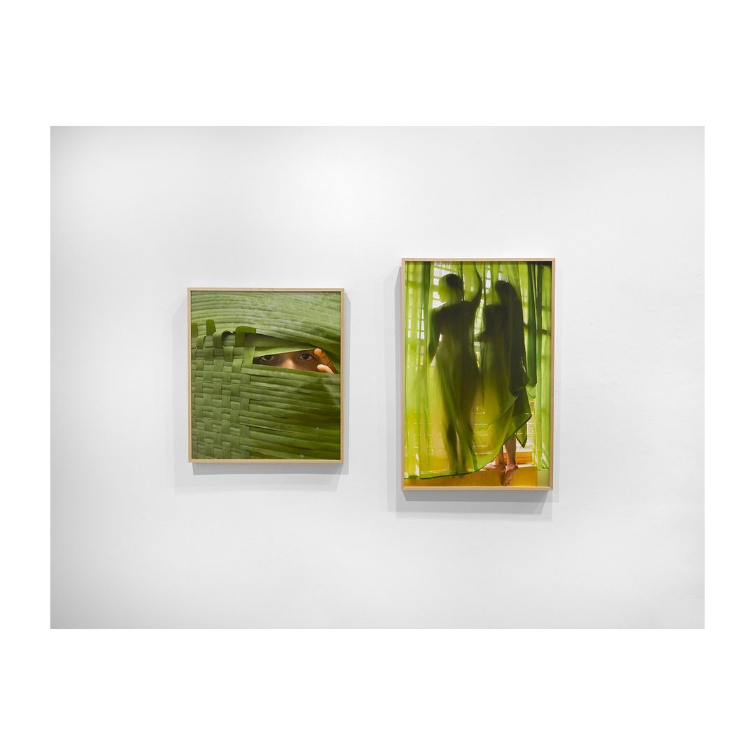 Two framed artworks hang on a white wall: one featuring a woven green surface with an eye peeking through, the other depicting two figures.