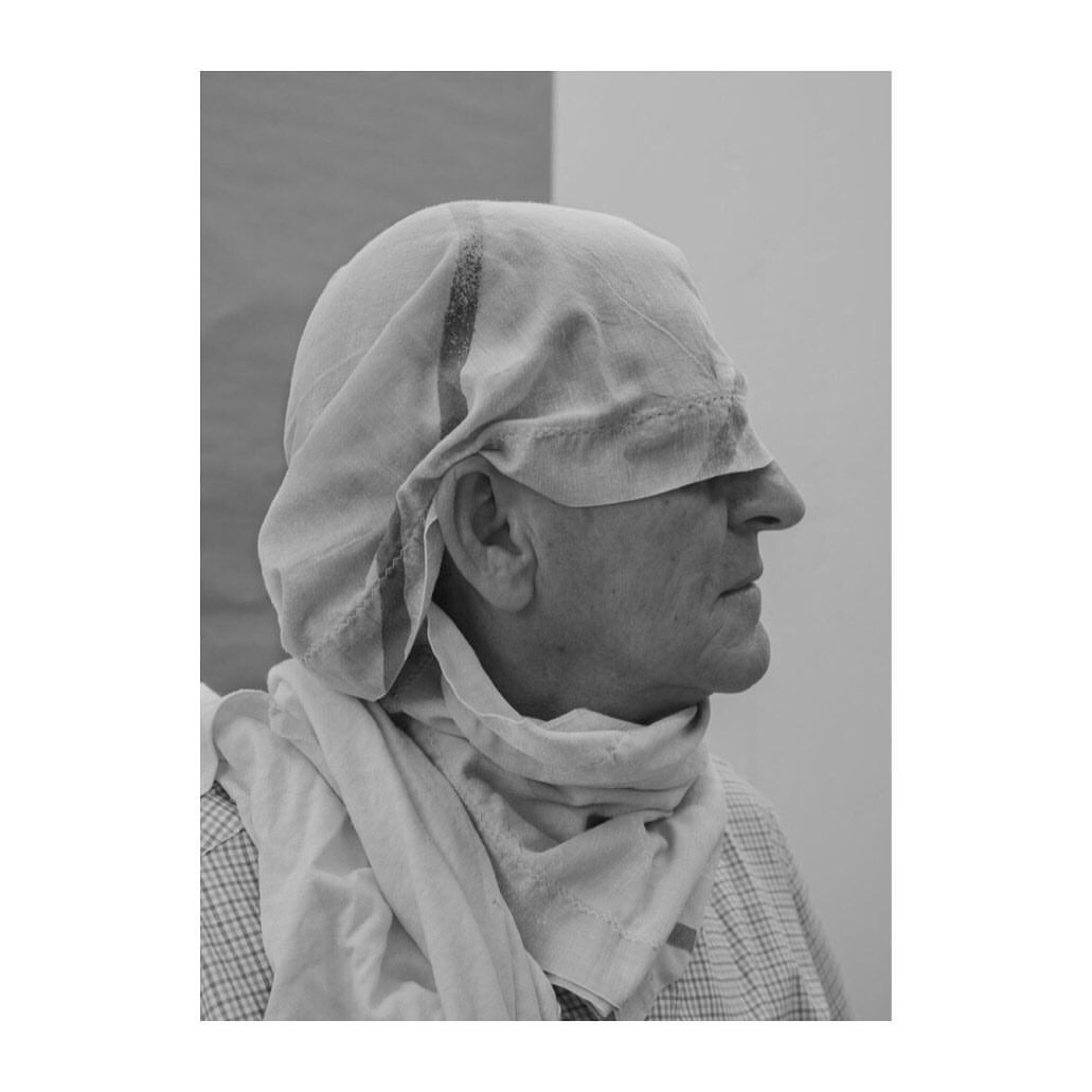 Black and white portrait of a mature adult with a cloth draped over their head and shoulders, in an indoor setting with neutral colors.