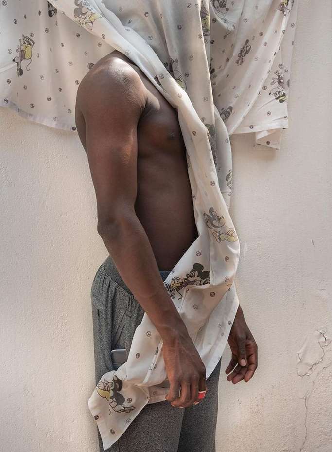 A shirtless adult man stands covered partially by a white sheet patterned with Mickey Mouse, against a light wall and shadow.
