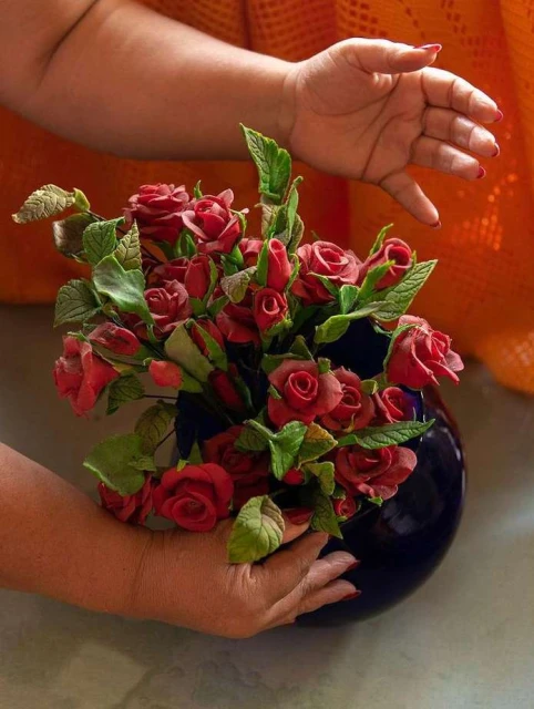 Close-up of a person holding a deep blue vase with red roses and green leaves, hands are partially visible. Orange background.