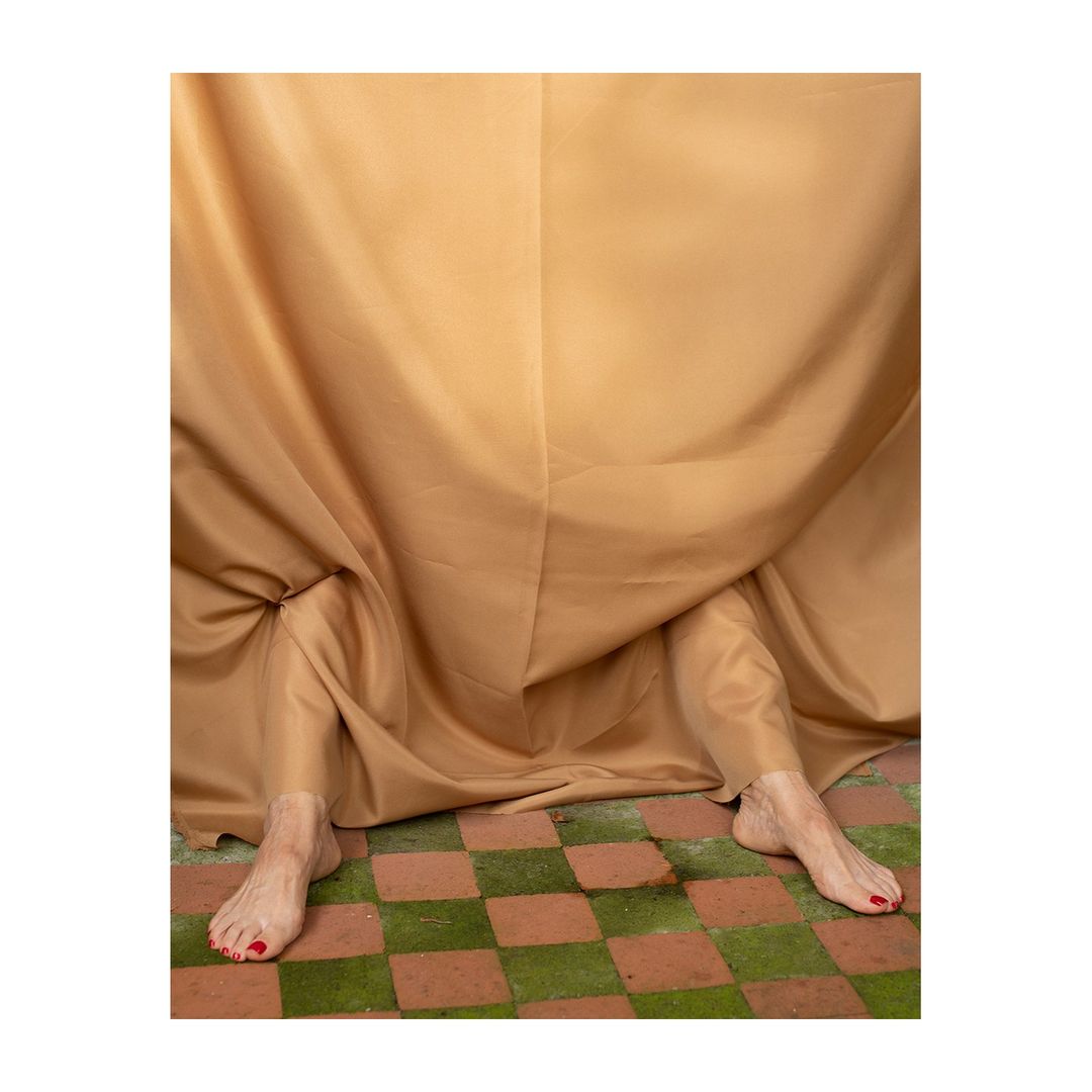 Abstract composition featuring bare legs extending from beneath a draped beige fabric on a checkered green and brown tile floor.