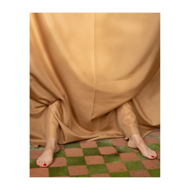 Abstract composition featuring bare legs extending from beneath a draped beige fabric on a checkered green and brown tile floor.