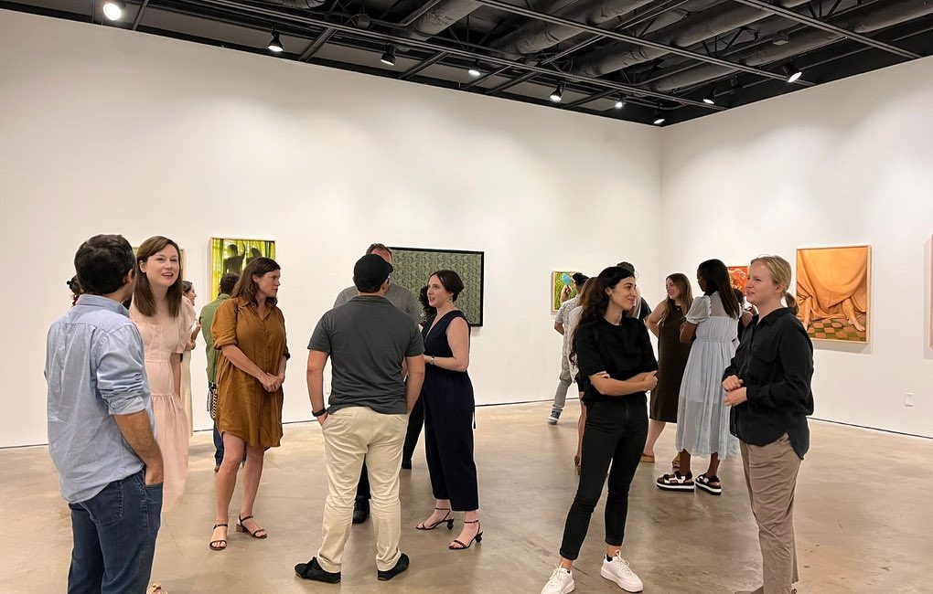 People attend an art gallery opening, viewing framed paintings in the white-walled space with recessed lighting and a polished concrete floor.