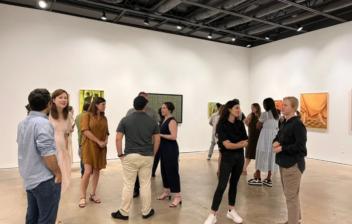 People attend an art gallery opening, viewing framed paintings in the white-walled space with recessed lighting and a polished concrete floor.