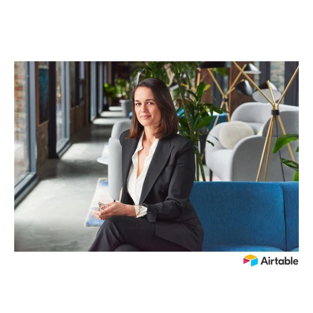 Professional headshot of a woman in a black business suit sitting on a blue couch, indoors in a modern office with natural lighting.