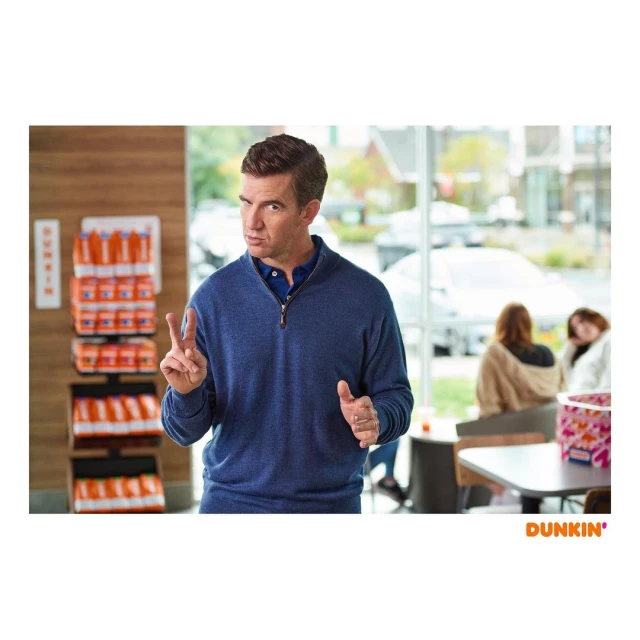Adult male in a blue quarter-zip sweater stands in a Dunkin' Donuts, holding up two fingers. Two women sit at a table in the background.