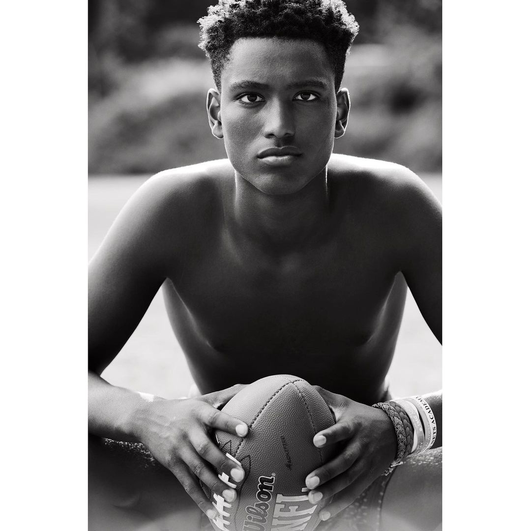 Black and white close-up of a shirtless young adult male holding a Wilson NFL football outdoors, surrounded by blurred greenery.