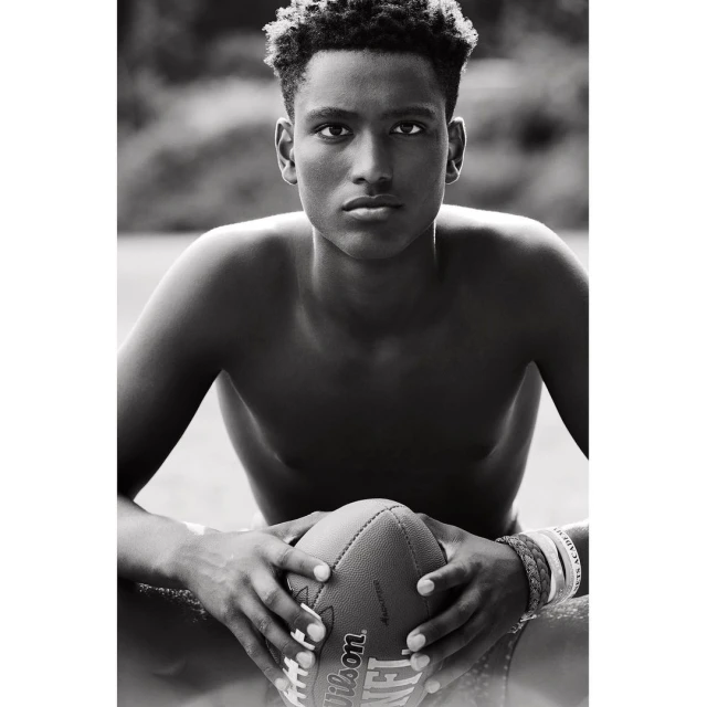 Black and white close-up of a shirtless young adult male holding a Wilson NFL football outdoors, surrounded by blurred greenery.