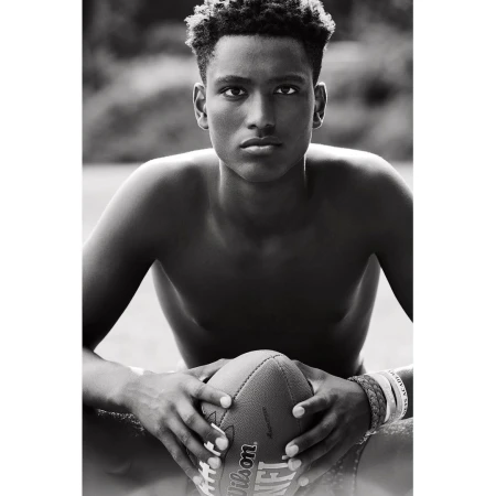Black and white close-up of a shirtless young adult male holding a Wilson NFL football outdoors, surrounded by blurred greenery.