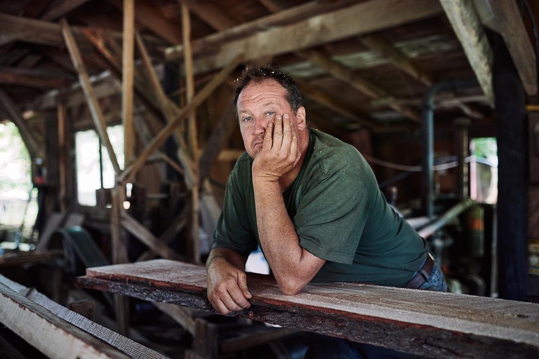 Adult male resting on wooden planks inside of old sawmill with rustic details and daylight.  Green shirt and relaxed pose. 