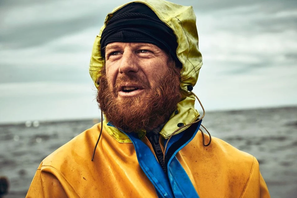 Close-up portrait of a red-bearded adult male wearing a yellow hooded rain jacket, looking up and to the left against an overcast sky.