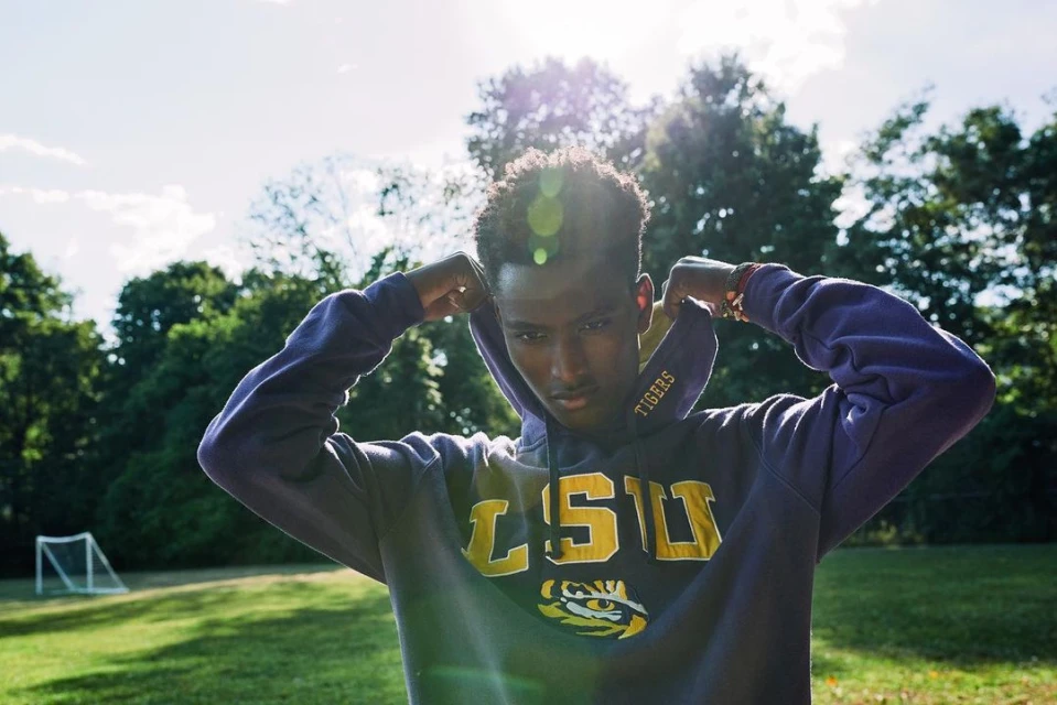 Young Black man wearing a purple LSU hoodie standing on a grassy field with trees, bright sunshine, and a goal in the background.