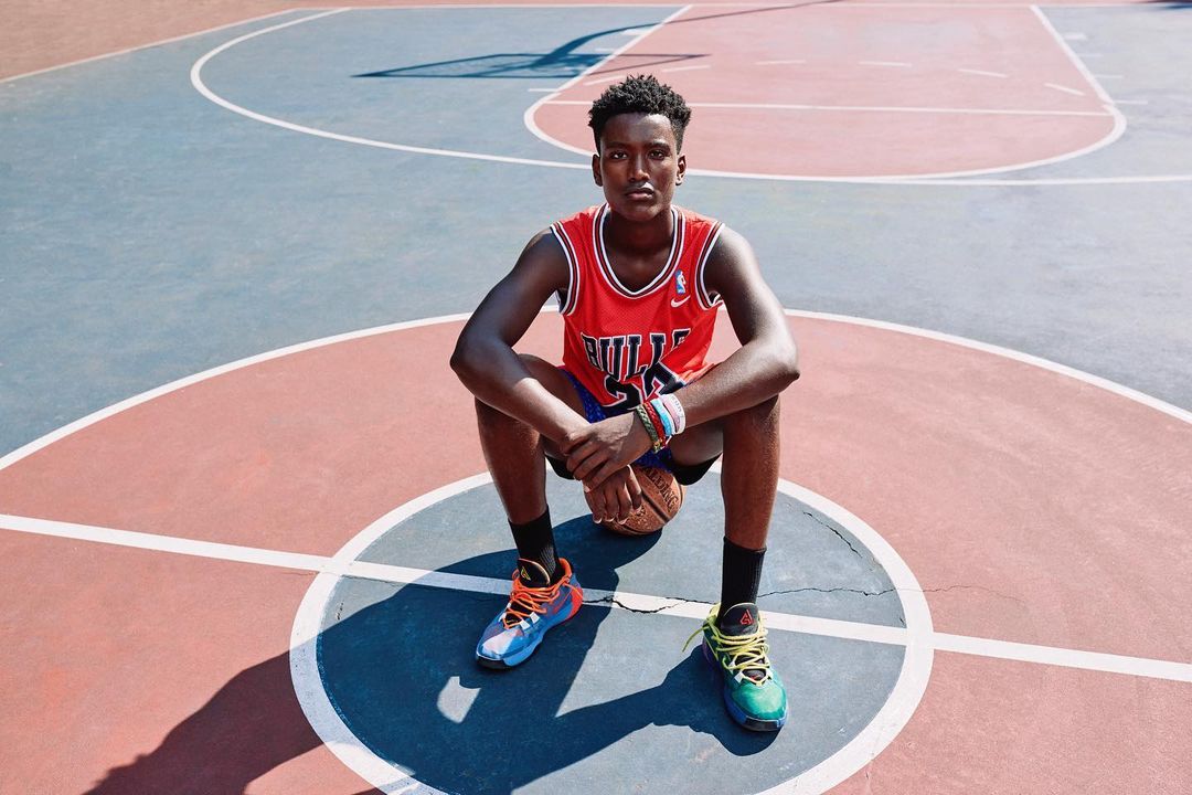 Young adult in a red Chicago Bulls jersey sits on a basketball, centered on an outdoor court with colorful basketball shoes.