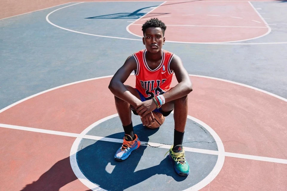 Young adult in a red Chicago Bulls jersey sits on a basketball, centered on an outdoor court with colorful basketball shoes.