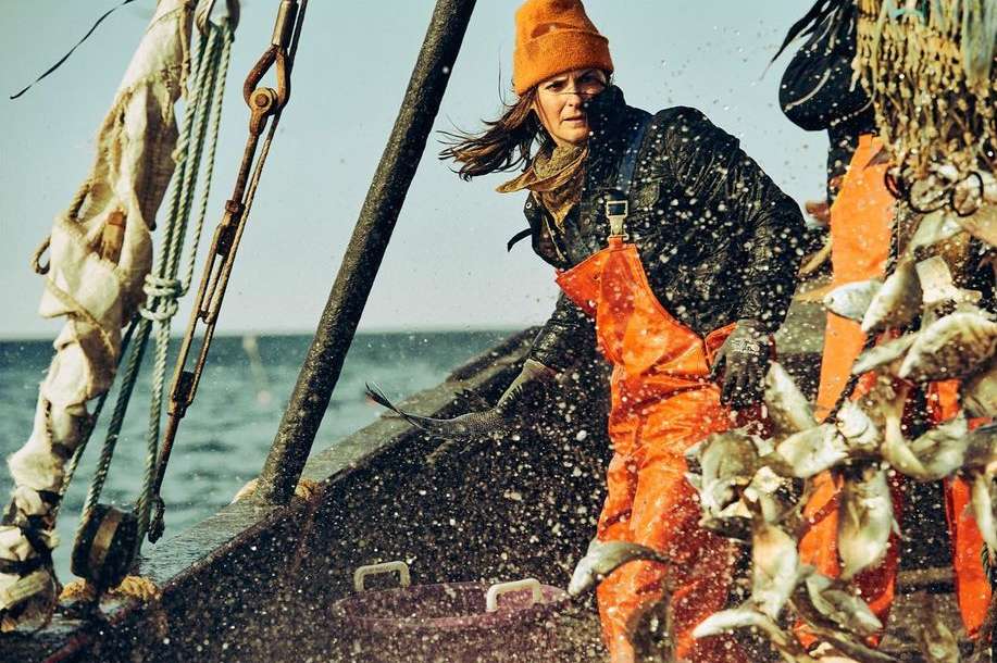 A woman in an orange hat and overalls stands on a fishing boat deck with a catch of fish and ocean water spraying around her.