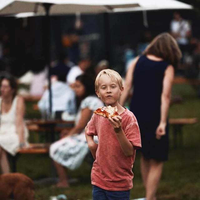 A blond boy in a red shirt eats pizza at an outdoor gathering, with other people and tables blurred in the background on the grass.